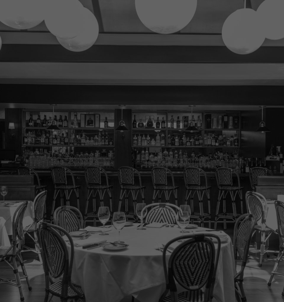 Empty restaurant with round tables set with wine glasses, plates, and utensils, and a bar in the background with bottles of alcohol on shelves.