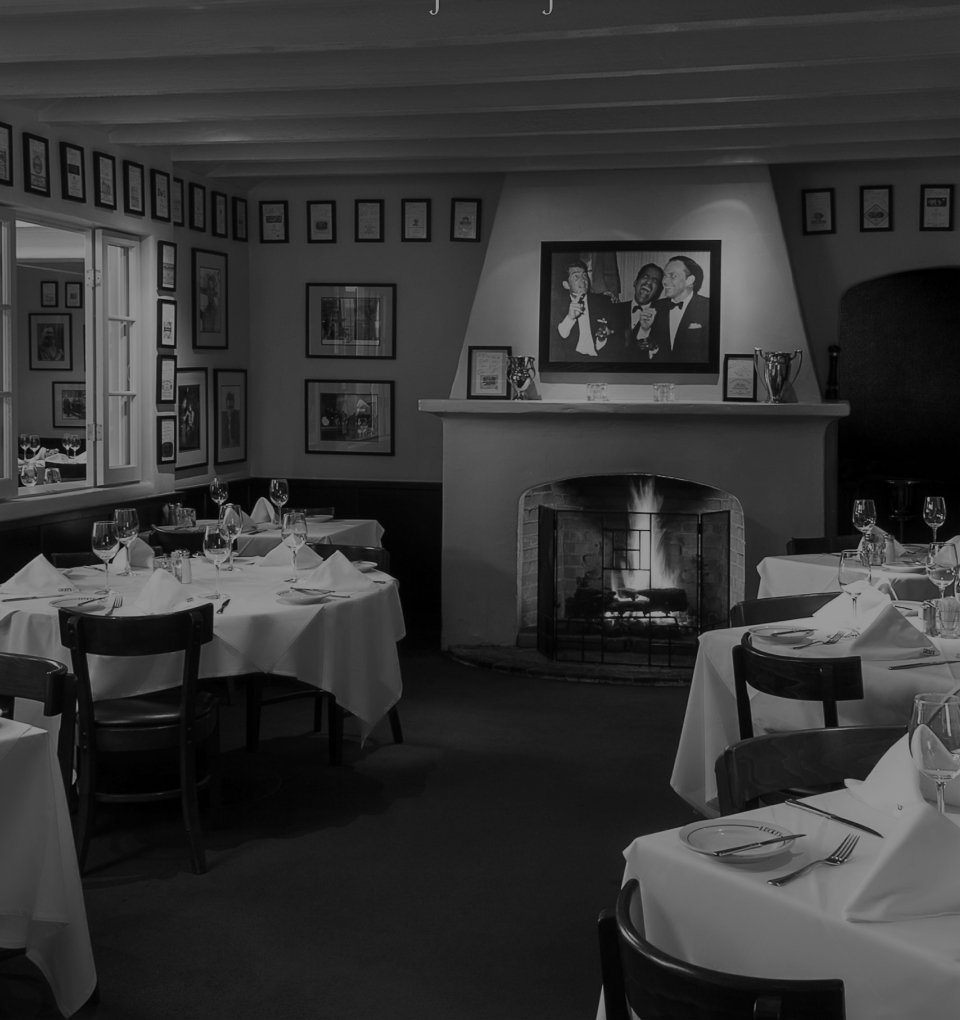Black and white photo of a cozy restaurant interior with tables set with white tablecloths, glasses, and napkins. There is a fireplace with a framed photo above it and walls decorated with various framed pictures.