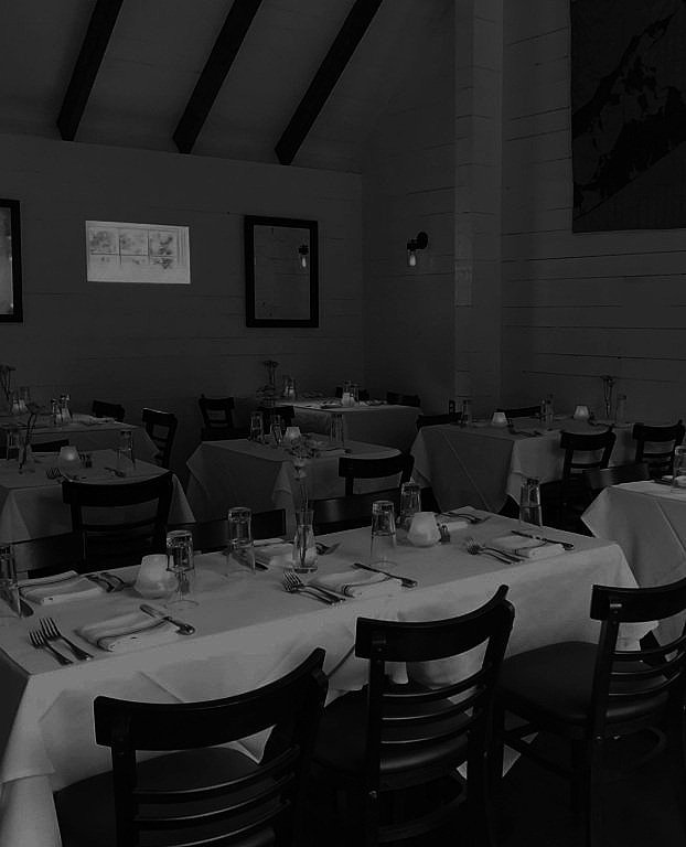 Empty restaurant dining room with tables set with white tablecloths, glasses, plates, and cutlery, illuminated by wall-mounted light fixtures.