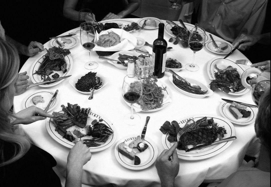 A black and white photo of a round table with seven people dining, with various plates of food, wine glasses, a bottle of wine, and side dishes.